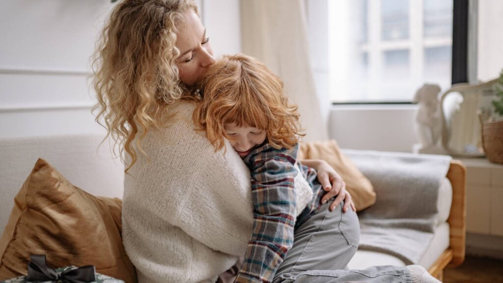 Press Centre Enquiries A mother gently cuddling her young child, holding them close against her chest with a warm, loving expression while the child rests peacefully in her arms.