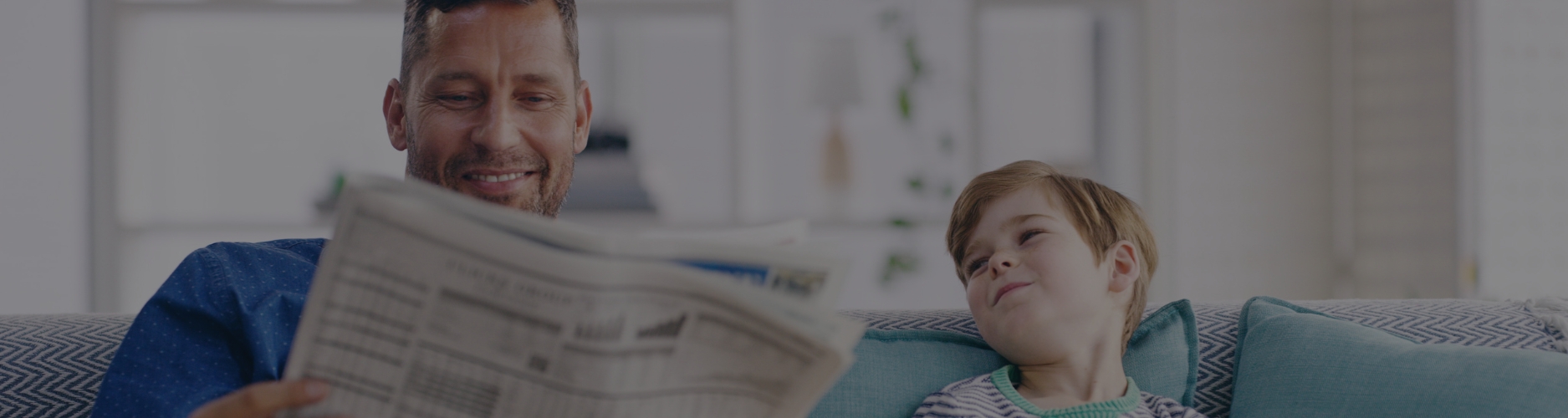 A father sitting on a sofa reading a newspaper while a smiling child looks up at him, sitting nearby and appearing happy and engaged in the quiet moment together.