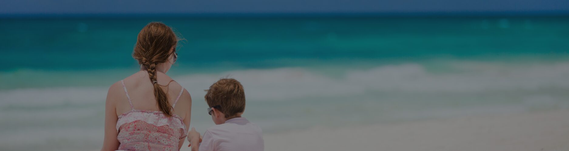 Teenage girl and younger brother sitting together on a beach looking out to the sea during the summer holidays.